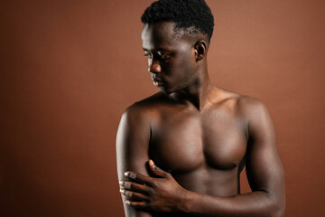 Confident African American young male with muscular naked torso standing in trendy jeans with hand on waist on brown background in studio and looking at camera