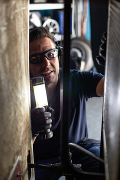 Professional male mechanic in protective glasses and headphones using flashlight while checking  details of machine during repair works in workshop