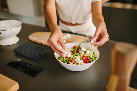Unrecognizable female adding Feta cheese in fresh vegetable salad while preparing healthy nutritious lunch in kitchen