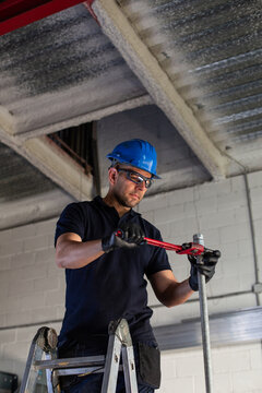 Low angle of cheerful male worker in hardhat and protective glasses standing on ladder and installing metal pipe with plumber wrench while working in workshop