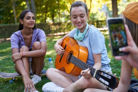 Cheerful multiracial female teenagers playing guitar and taking photo on mobile phone while sitting on green lawn in park and enjoying summer day together