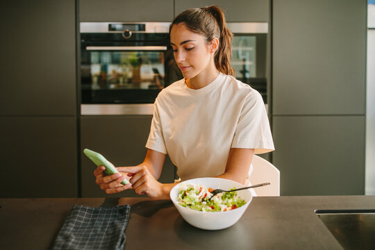 Peaceful female sitting at table in kitchen and eating tasty vegetable salad while reading news on social media on smartphone
