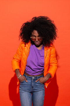 Young black female in purple shirt with denim and orange jacket standing with hands in pockets on red background looking down