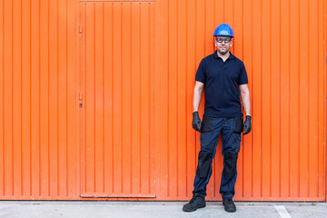 Positive confident adult male workman in blue protective hardhat and eyeglasses while standing against bright orange wall of workshop