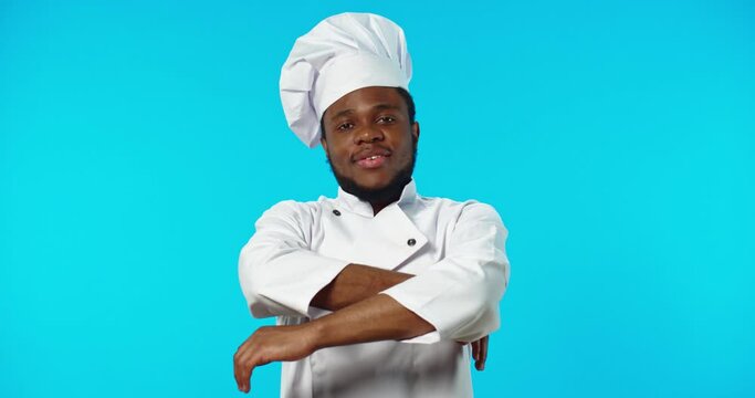 Portrait of joyful happy African American handsome bearded young man in white chef uniform and hat standing isolated on blue background in studio looking at camera in good mood, cook concept