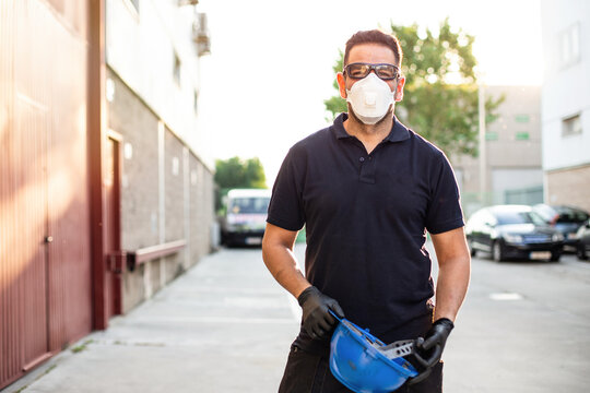 Adult male mechanic in protective glasses and respirator holding blue hardhat in hands and looking at camera while standing near workshop building on street
