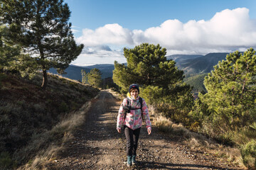 Young and attractive female with a colourful jacket hiking in a route between pine trees and other plants with a digital camera.