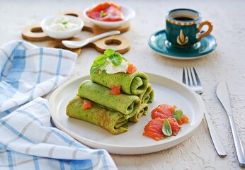 Spinach pancakes rolled up on a white plate on a light concrete background. Served with salted salmon and soft cheese.