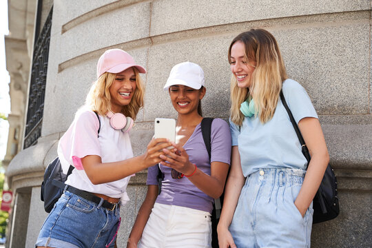 Cheerful young multiracial girlfriends in casual outfits using smartphone while standing together near stone building in city in summer day