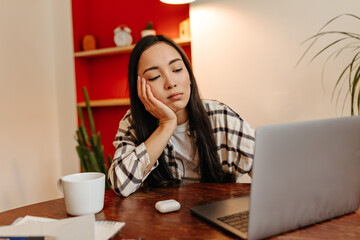 Asian girl in shirt leaned on desktop and tiredly looking at laptop