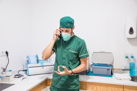 Male dentist in mask and uniform speaking on smartphone while holding dental cast in modern dental hospital