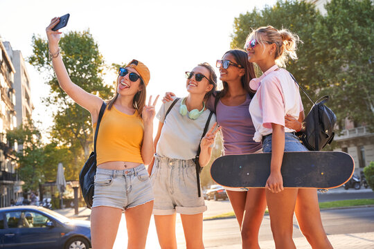 Cheerful trendy young multiracial girlfriends in sunglasses with backpacks and skateboard taking selfie on mobile phone while gathering together on urban square in summer day