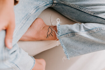 High angle of unrecognizable female in jeans and with tattoo in shape of sun on leg sitting on sofa