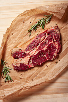Top view of fresh uncooked juicy rib eye beef steak with fresh rosemary and pepper placed on baking paper sheet on table