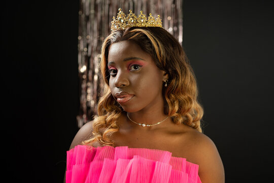 Self assured young black lady with long curly hair and makeup in pink fashionable dress and golden crown looking at camera during beauty contest