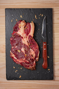Top view of fresh uncooked rib eye beef steak with rosemary sprig and dried spices on black slate board