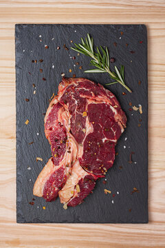 Top view of fresh uncooked rib eye beef steak with rosemary sprig and dried spices  on black slate board