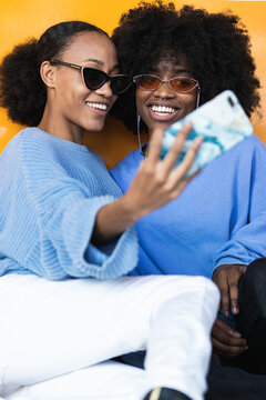 Cheerful young African American women in stylish sunglasses and lilac clothes taking selfie on mobile phone against yellow background