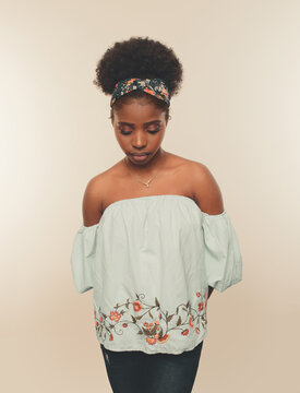 Young slim African American female model with Afro hair bun and headband wearing stylish blue crop top and jeans standing against beige background
