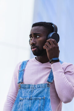 Cool African American male hipster in trendy outfit standing on street and listening to music in wireless headphones