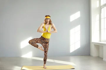 Deurstickers Weird fit guy in hilarious leopard leggings standing on gym mat and doing Tree pose. Funny young man in retro activewear practicing Vrikshasana yoga exercise at home. Balance and harmony humor concept © Studio Romantic