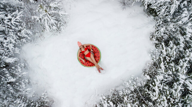 A girl in a red swimsuit lies on an inflatable circle in the shape of a watermelon. Snow, winter, cold. Shooting from above.