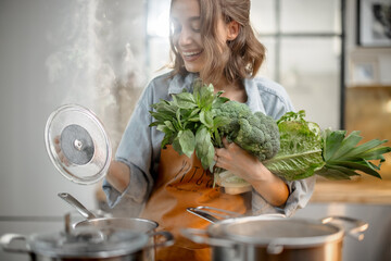 Pretty housewife in apron with fresh vegetables near cooker with boiling pans on the kitchen. Healthy green cooking concept. High quality photo