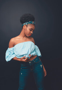 Young slim African American female model with Afro hair bun and headband wearing stylish blue crop top and jeans standing against black background