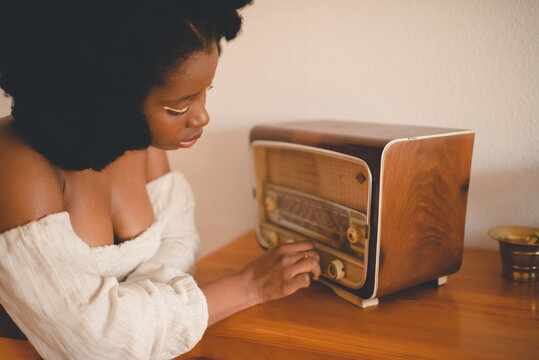Side view of young African American female sitting at table and tuning old fashioned radio receiver while resting at home