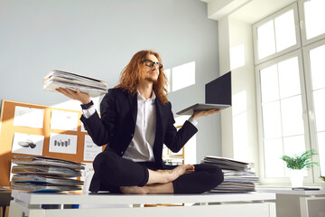 Calm barefoot busy office worker in suit sitting in lotus yoga pose on desk with papers, holding paperwork pile and laptop, breathing deeply, meditating and relaxing. Balance and stress relief at work