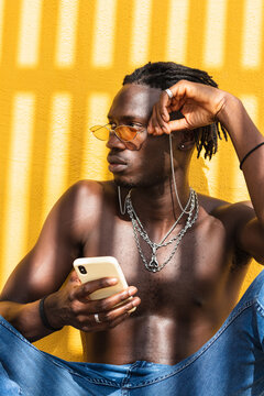 Serious Young Shirtless African American Man In Trendy Sunglasses And With Metal Chains Browsing Mobile Phone While Sitting Against Yellow Wall With Striped Shadow