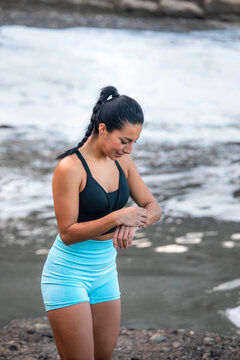 High angle of content sportswoman standing on seashore and checking pulse on modern smart watch during active workout in summer