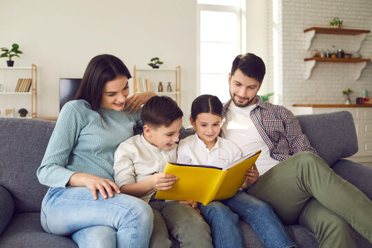 Young parents with little daughter and son read fairy tales and look at pictures in a book sitting on the couch at home. Father, mother and their children are smiling while spending time together.