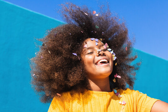 Low angle of happy carefree teen African American girl with long curly hair and colorful confetti on face having fun against blue wall in sunny summer day