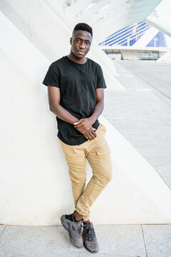 Masculine African American male in casual style clothes standing with crossed legs and leaning on building in city while looking at camera