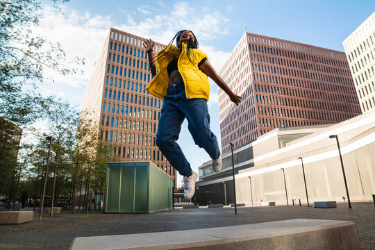 Happy black woman in trendy clothes laughing and leaping up on street of modern city looking at camera