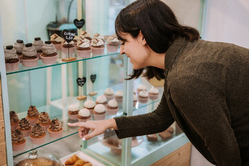 Side view of happy young female customer pointing at cupcake on display of confectionery shop while choosing sweets for purchase