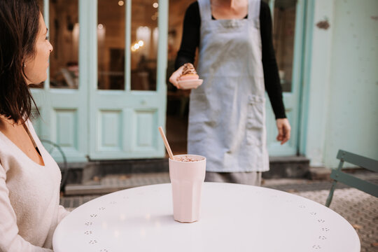 Crop waitress serving cupcake to happy female customer sitting at table with glass of frappe coffee in cozy confectionery cafe