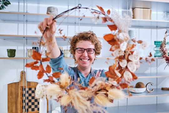 Positive female florist standing in flower salon and showing creative wreath made of dried leaves and plants