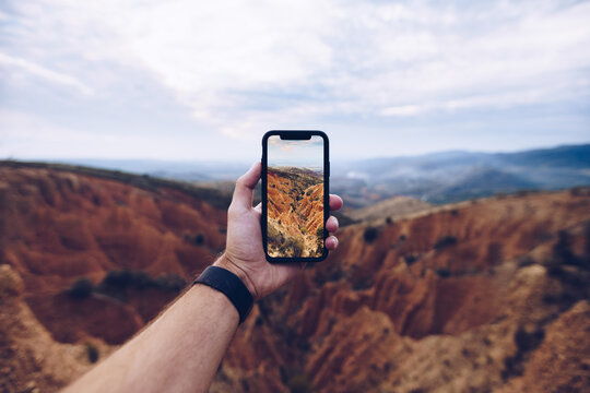 Crop unrecognizable male hiker shooting picture of eroded mountainous landscape on mobile phone while travelling through highlands and exploring wild nature