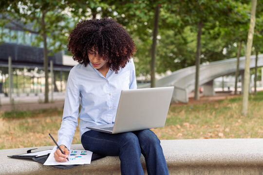 Focused female entrepreneur sitting on stone bench in park and working remotely while on laptop and taking notes in documents