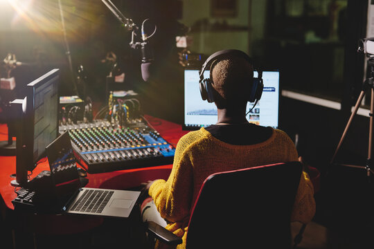Back view of ethnic female in headphones sitting at table with mixing console and speaking in microphone while working in broadcast studio