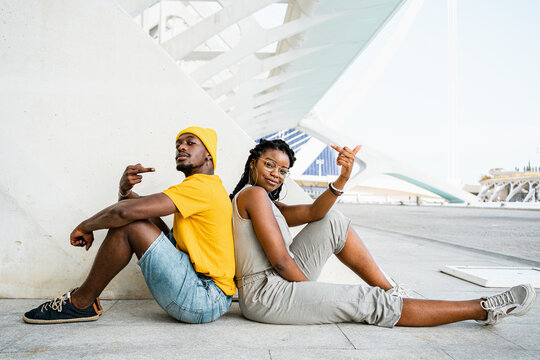 Side view of young stylish African American friends sitting back to back on street and showing middle fingers while looking at camera