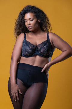 Attractive African American overweight female model with long curly hair wearing delicate lace bra looking down against yellow background