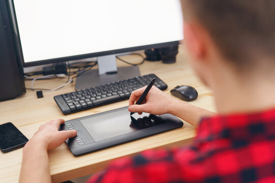 Close-up, a young adult guy using a pen tablet does retouching in front of a computer at home. The photographer processes the photos. A blank white computer screen. Freelancer. Remote work. Blogging.