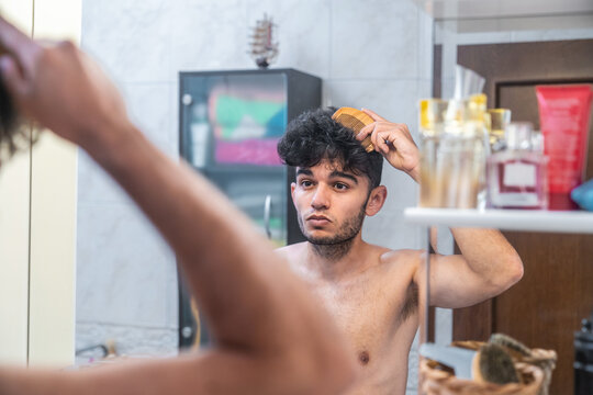 Young shirtless bearded male with stylish haircut brushing hair with comb and looking at mirror during grooming routine in bathroom at home