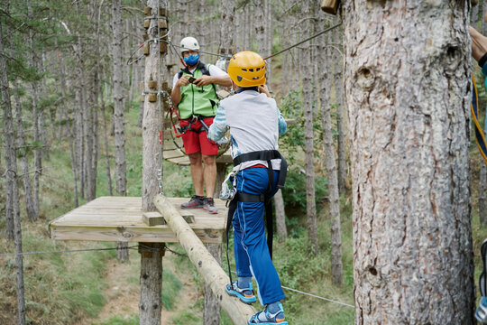 Unrecognizable Boy In Helmet Using Safety Rope To Walk On Log Towards Man While Climbing Obstacle Course In Adventure Park In Forest