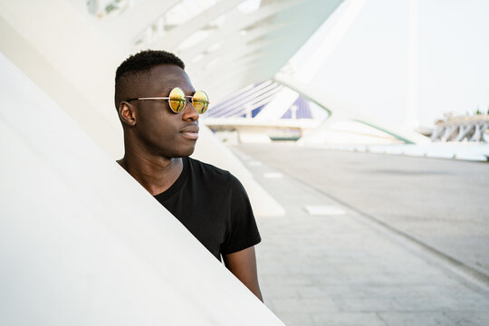 Young African American male wearing trendy round sunglasses standing in urban environment and confidently looking away