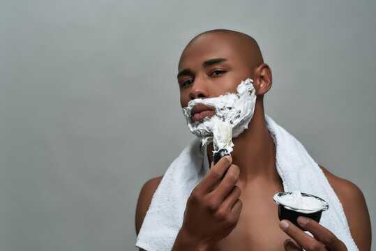 Shirtless attractive young african american man looking at camera, using brush while applying shaving foam on his face, posing isolated over gray background