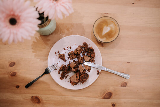 Top View Of Plate With Crumbled Chocolate Dessert Served With Glass Cup Of Aromatic Latte On Wooden Table With Flowers In Cozy Cafeteria